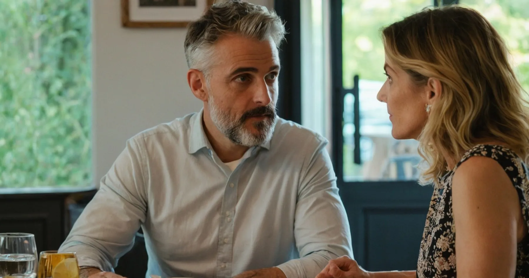 Un homme à la barbe grise et une femme blonde, de profil, discutent sérieusement à une table avec des verres